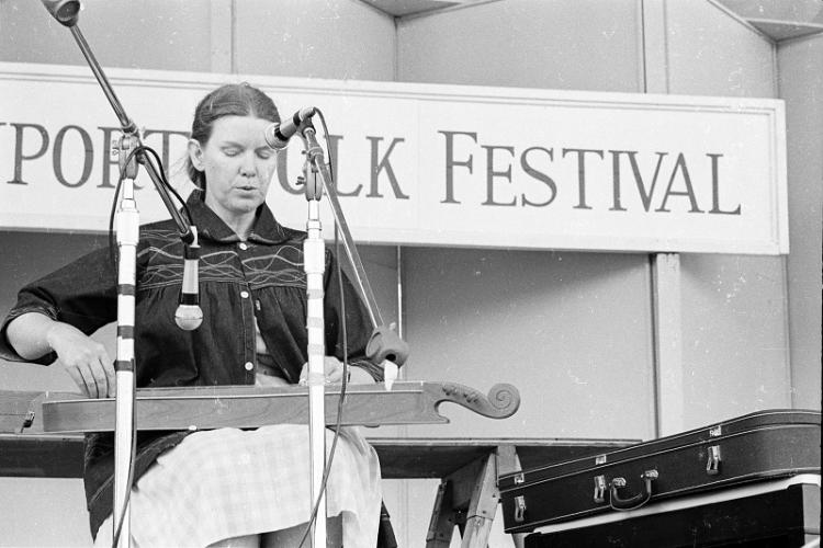 Jean Ritchie sitting on stage behind two microphones, singing while playing a dulcimer placed on her lap.