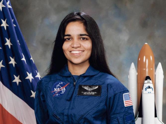 Kalpana Chawla in her blue NASA uniform stands in front of an American flag and a model of a space shuttle