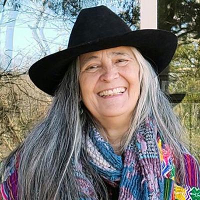 Color headshot photograph of a woman with long gray hair wearing a black hat and purple and blue patterned neck scarf with trees and grass in the background.