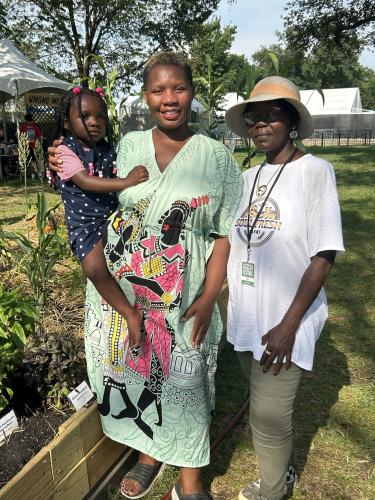 Two African American adult women stand next to a raised garden bed.  The younger woman holds a child and wears a green dress printed with stylized figures.  The woman standing next to her wears a hat and t-shirt with the logo for Soul Fresh Farms.