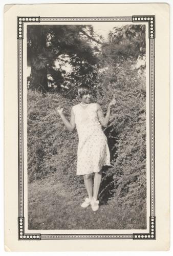 Black and white photograph of a young black woman wearing light-colored printed dress and white shoes with bows at the ankle stands in front of bushes that form a hedge. Long-needle pine trees are behind the hedge. The photograph is mounted on a card and surrounded by a printed frame.   