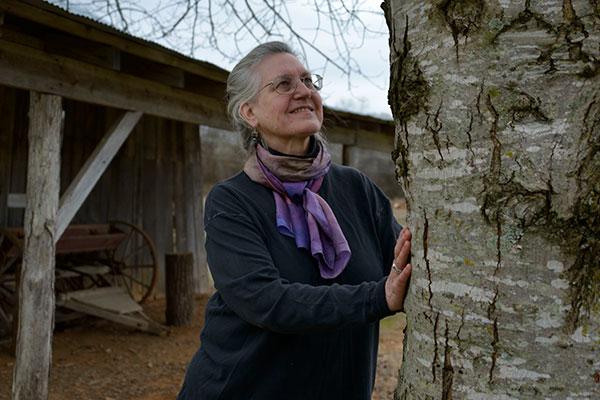 Photograph of a woman with gray hair, wearing glasses, a purple-toned neck scarf, and dark clothing looking at a large tree with both of her hands on the trunk.  A wood shelter with the wheel and tongue of a horse-drawn wagon visible is in the background. 