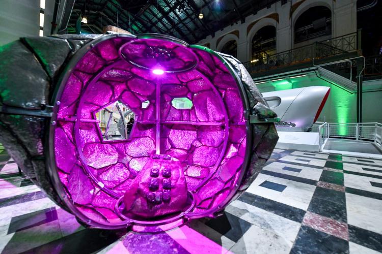  Pod-like steel artwork with blobby circular pattern within, lit in purple light. It is on display in a museum gallery.