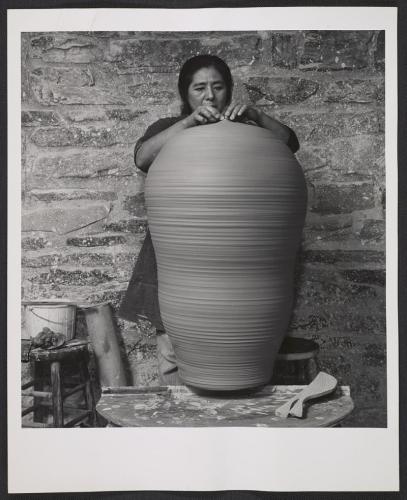 Toshiko Takaezu stands in a studio behind a large cylindrical clay vessel, finishing the very top. The clay artwork comes up to her chin.