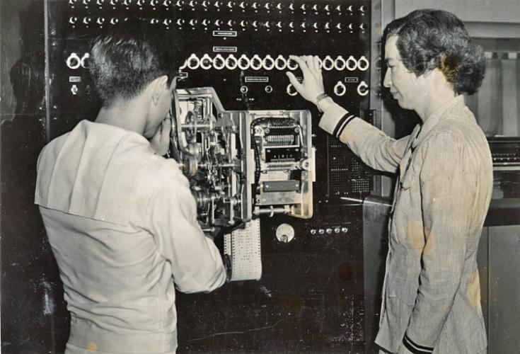 A young navy ensign stands with Grace Hopper, wearing her navy uniform and standing in profile.  She has her right hand on one of the many dials that run across the top of the Mark I computer.