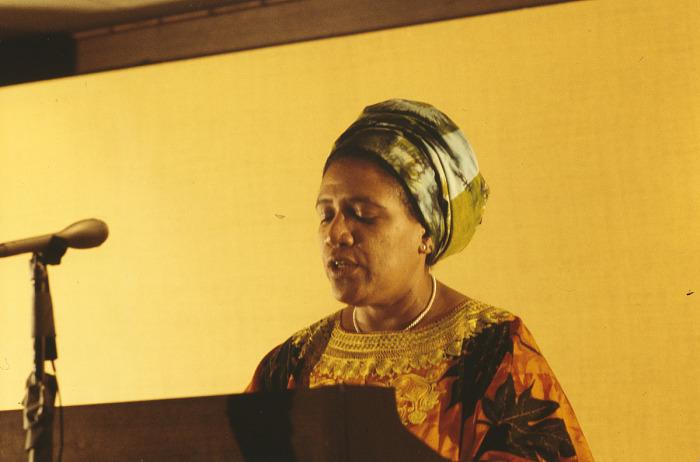 Candid color photo of Audre Lorde from the shoulders up standing at a microphone and podium.   