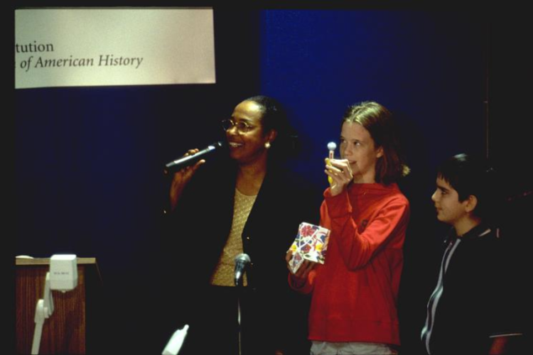 Dr. Bath holds a microphone, speaking from a stage. Two young students stand next to her.