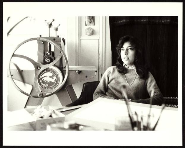 Black and white photo of a woman sitting at a desk. She has long wavy hair and wears a sweater. There is drawing-like equipment in front of her, including a mechanical device.