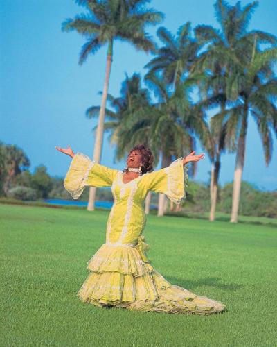 Celia Cruz stands outside on manicured grass with her arm stretched wide. She wears a bright yellow bata cubana (Cuban rumba dress) with wide bell sleeves. Her mouth is open, as if she is singing or shouting. There are palm trees in the background.