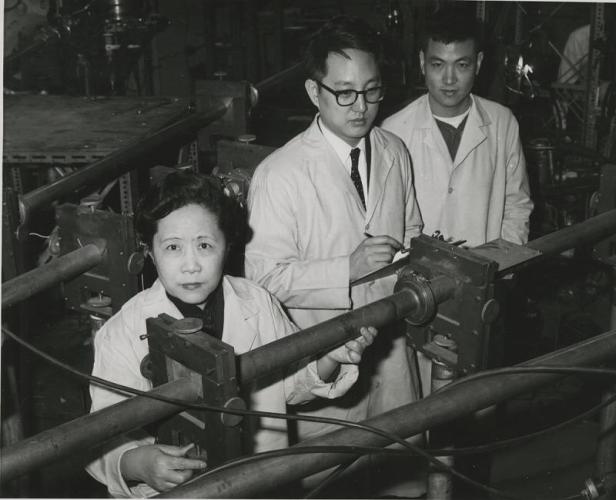 Chien-shiung Wu, Y.K. Lee, and L.W. Mo. wear lab coats and stand in front of two long, horizontal tubes that strech out of frame.