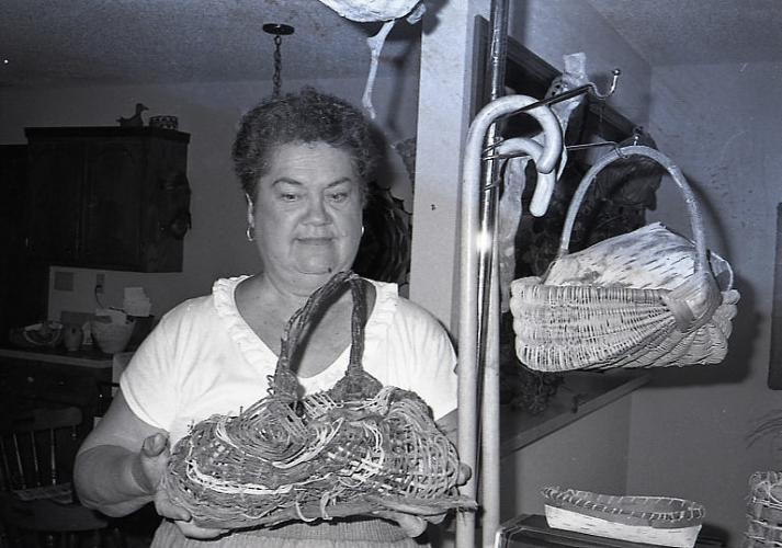 Black and white photo of Grace Thorpe holding a woven basket in a room with two canes and several other woven baskets.