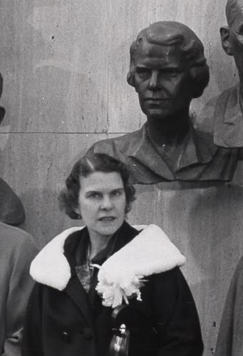 Black and white photograph of a woman standing in front of her own portrait bust wearing a dark coat with a white collar.