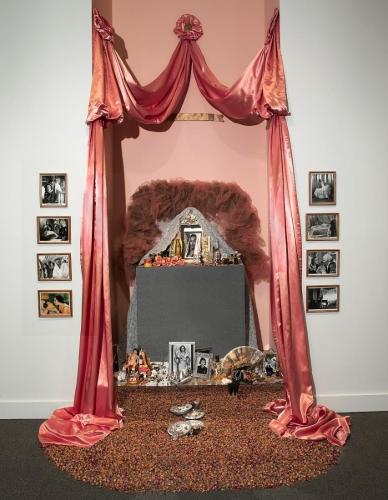 Color photograph of installation art in the form of an ofrenda.  An altar formed by coral pink drapery surrounds a makeup vanity table with a white lace and pink fabric crown. The altar is flanked by four movie stills of Dolores del Rio on each side in a vertical configuration. The vanity table holds black and white photographs, candles, souvenirs, images of the Virgin of Guadalupe and examples of Mexican folk art. Miniature, dried pink rosebuds spill out of the ofrenda onto the floor.