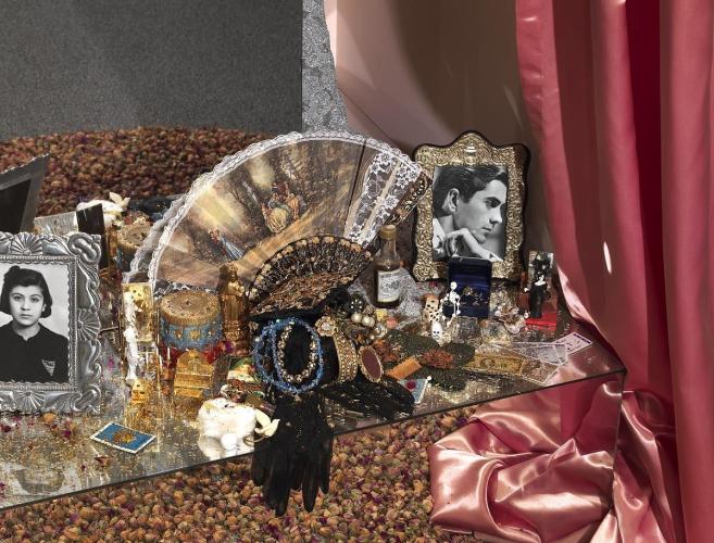 Color photograph of a collection of objects including portrait photos, jewelry, gloves, an open fan with a painting printed on the fabric, and dice on top of a mirrored surface that reflects a pile of dried pink rosebuds. 