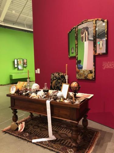 Color photograph of a museum gallery. In the right foreground, against a pomegranate-red wall there is an installation with a tri-fold mirror on the wall, desk, chair, and rug. On the desk there are candlesticks, a globe, seashells, photos, an hourglass, and a book with a skull resting on top of it. In the left background there is a lime green wall with a tri-fold mirror and horizontal exhibit case.  