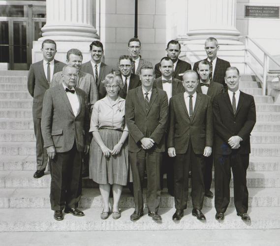 Black and white photograph of one woman and thirteen men in formal attire standing in three rows on the white front steps of the National Museum of Natural History.  