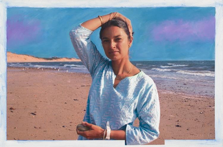 Jhumpa Lahiri stands on a beach and looks, squints as she looks into the camera. Her right hand rests on her head, pulling back her hair. Her left hand holds a stone. She wears a loose-fitting top and bangle bracelets.