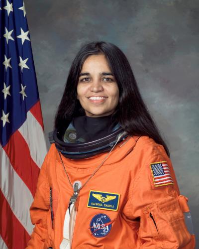 Color portrait of Kalpana Chawla in her orange NASA suit with the American flag behind her to the left.