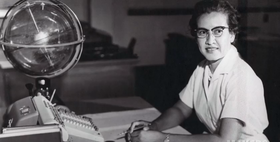 A black and white photo of Katherine Johnson sitting at a desk in front of a typewriter and open notebook.