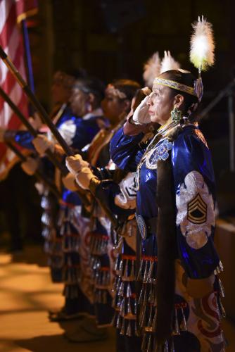 Mitchelene BigMan and a line of other Native American Women Warriors holds a flag. They all wear ceremonial dresses.