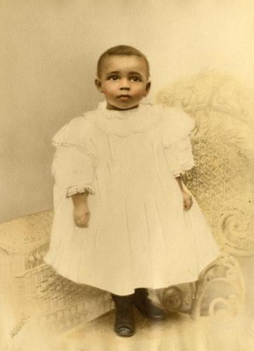 A toddler Marian Anderson looks up at the camera in this gelatin silver print. It is hand-colored with an illustrated quality.