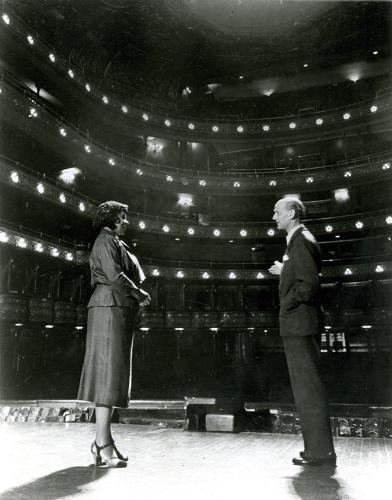 Marian Anderson and Sir Rudolf Bing stand on the stage of the Metropolitan Opera House, speaking to one another. The empty seats can be seen behind them.