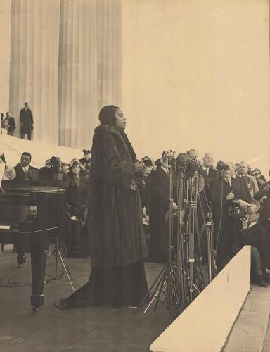 Marian Anderson sings on the steps of the Lincoln Memorial. She wears a large fur coat. Press and audience members can be seen to her left.