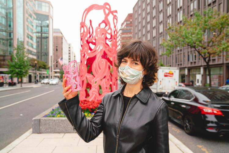 Rania Hassan stands in font of Marker, a tall metal sculpture depicting fluid pink strands intersecting. Hassan holds a model of the artwork in her hand.