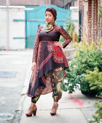 Woman wearing a fashionable ensemble of tunic, pants, headwrap, and neck scarf. The pants and neckscarf are made of a camoflauge print fabric. The tunic is made of an African wax-print fabric.