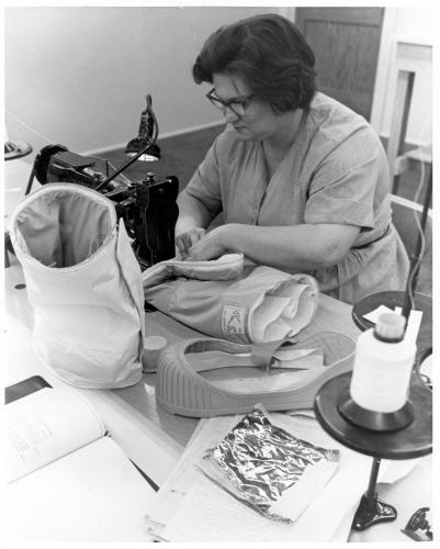 A woman wearing glasses sits at a dark-colored sewing machine surrounded by a leg piece standing upright on the table, part of a rubber sole, a reflective square of material, and an industrial spool of thread.