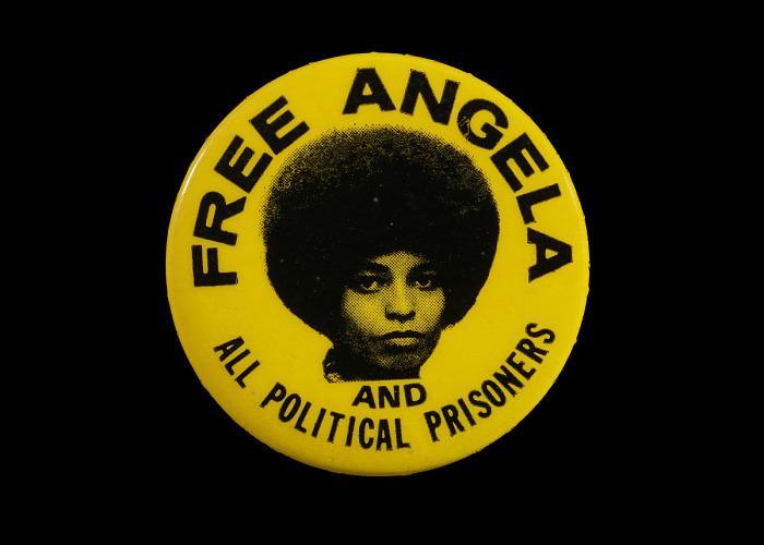 A round, yellow pinback button with a photographic portrait of Angela Davis in the center, pictured from the neck up. Button text: Free Angela / And / All Political Prisoners.