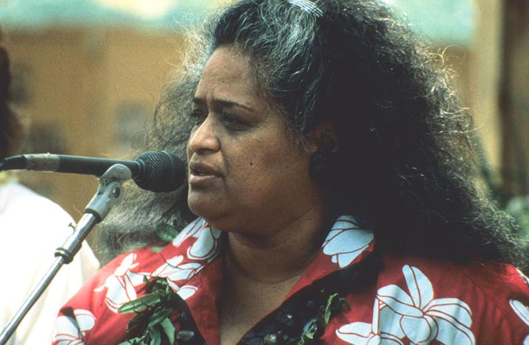 Pualani Kanakaʻole Kanahele sings into a microphone. She wears a lei and a red shirt with white flowers on it.