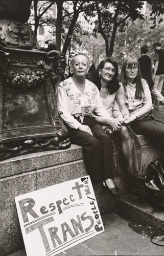 Sylvia Rivera, Christina Hayworth and Julia Murray sit on a ledge in a park in New York City. A sign near them reads: Respect Trans People/Men!