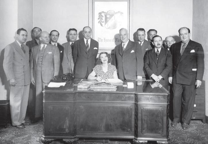 Tillie Lewis sits at an office desk. She is surrounded by men in suits.