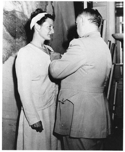 Virigina Hall looks at General Donovan as he pins the Distinguished Service Cross to her top. She wears a light colored suit with a skirt. Her hair is done in an updo with a wide light-colored hairband.