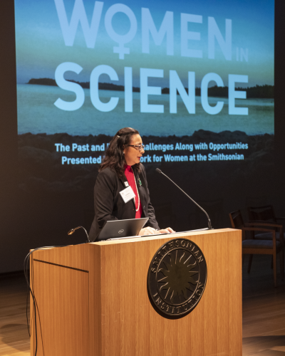 Oris Sanjur speaks at a podium. The words Women in Science are projected on a screen behind her.