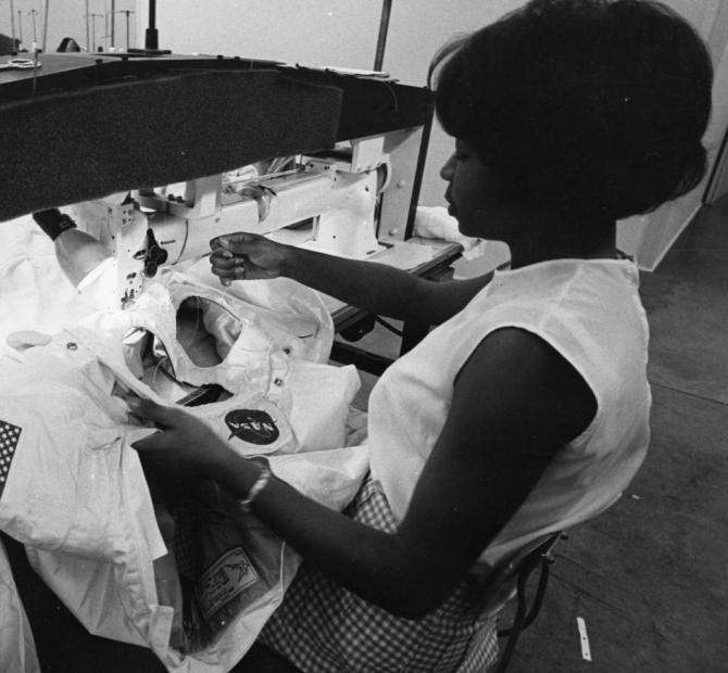 Black and white photograph of African American woman sitting at a long-arm sewing machine working on the iconic white outer layer of an Apollo spacesuit.